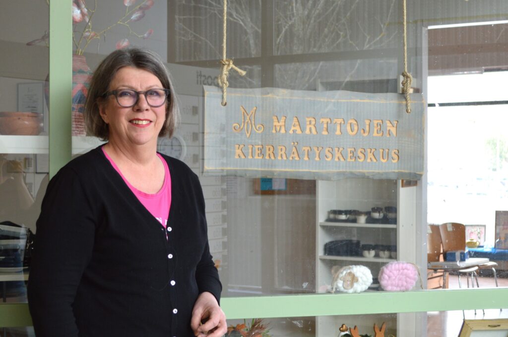 Nainen hymyilee ikkunan edessä. Ikkunassa on puinen kyltti, jossa lukee "Marttojen kierrätyskeskus".